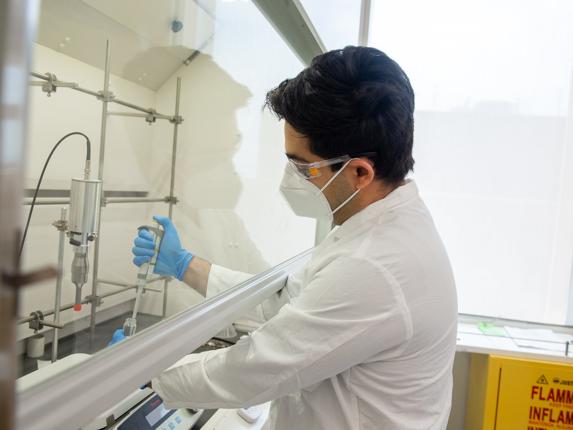 A man in a white lab coat, N95 mask and blue gloves uses a syringe-type device in a test tube.