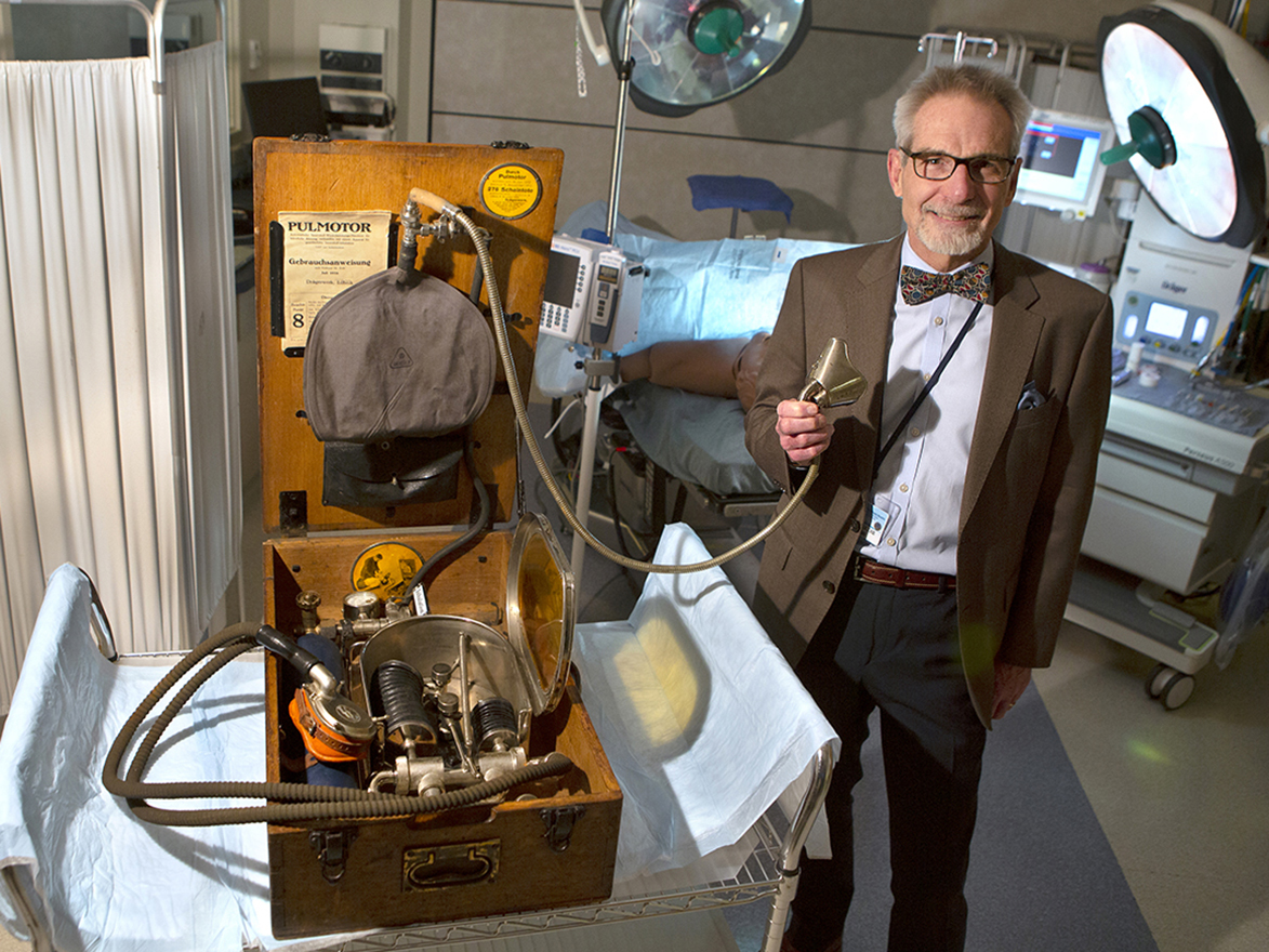 Man smiling, standing in an operating room, holding a face mask from an antique piece of anesthesiology equipment.