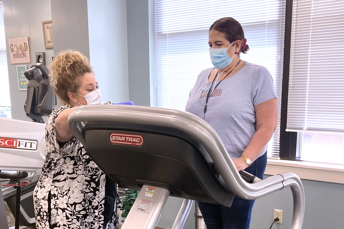 Two women are pictured in the photo. The one on the left is wearing a face mask and is leaning on the treadmill. The woman on the right is wearing a face mask and standing on the treadmill. 