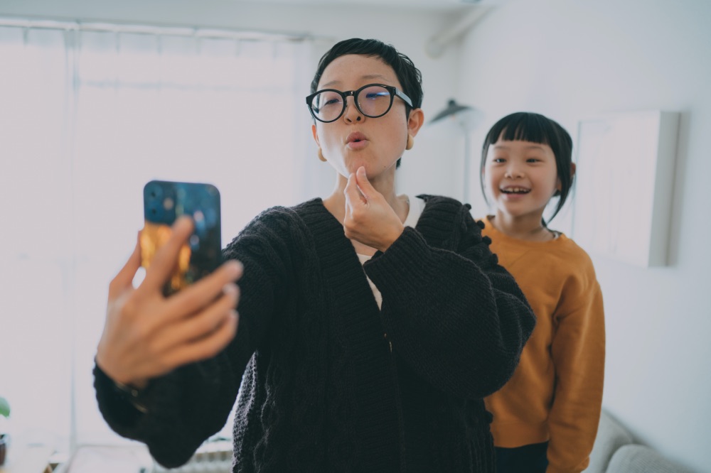 A woman uses sign language to communicate into a smartphone while a child behind her watches and smiles.