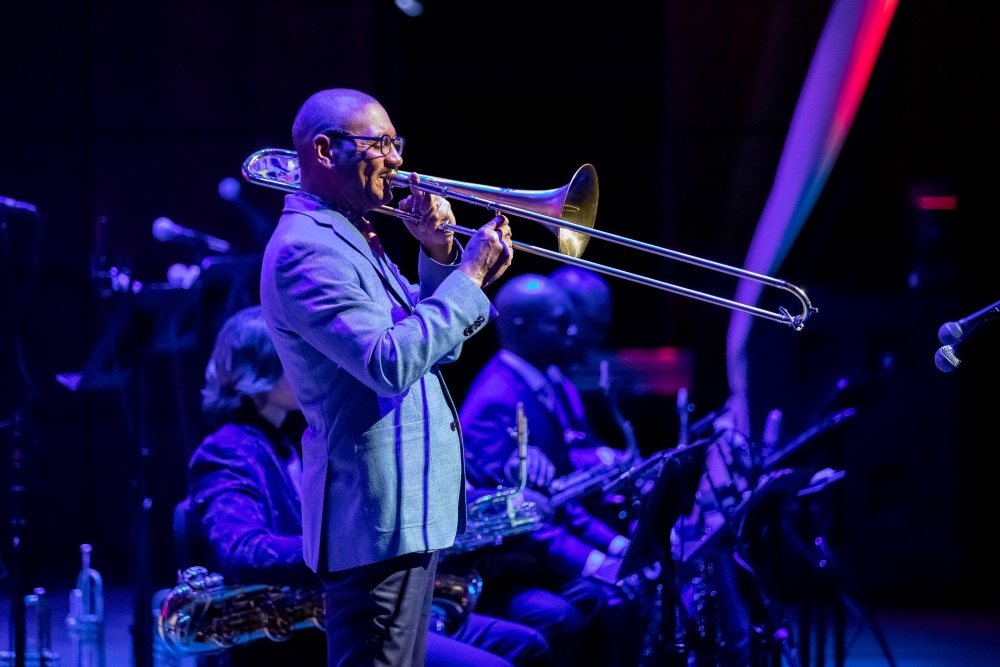 A man wearing eyeglasses and a suit stands and plays a trombone.