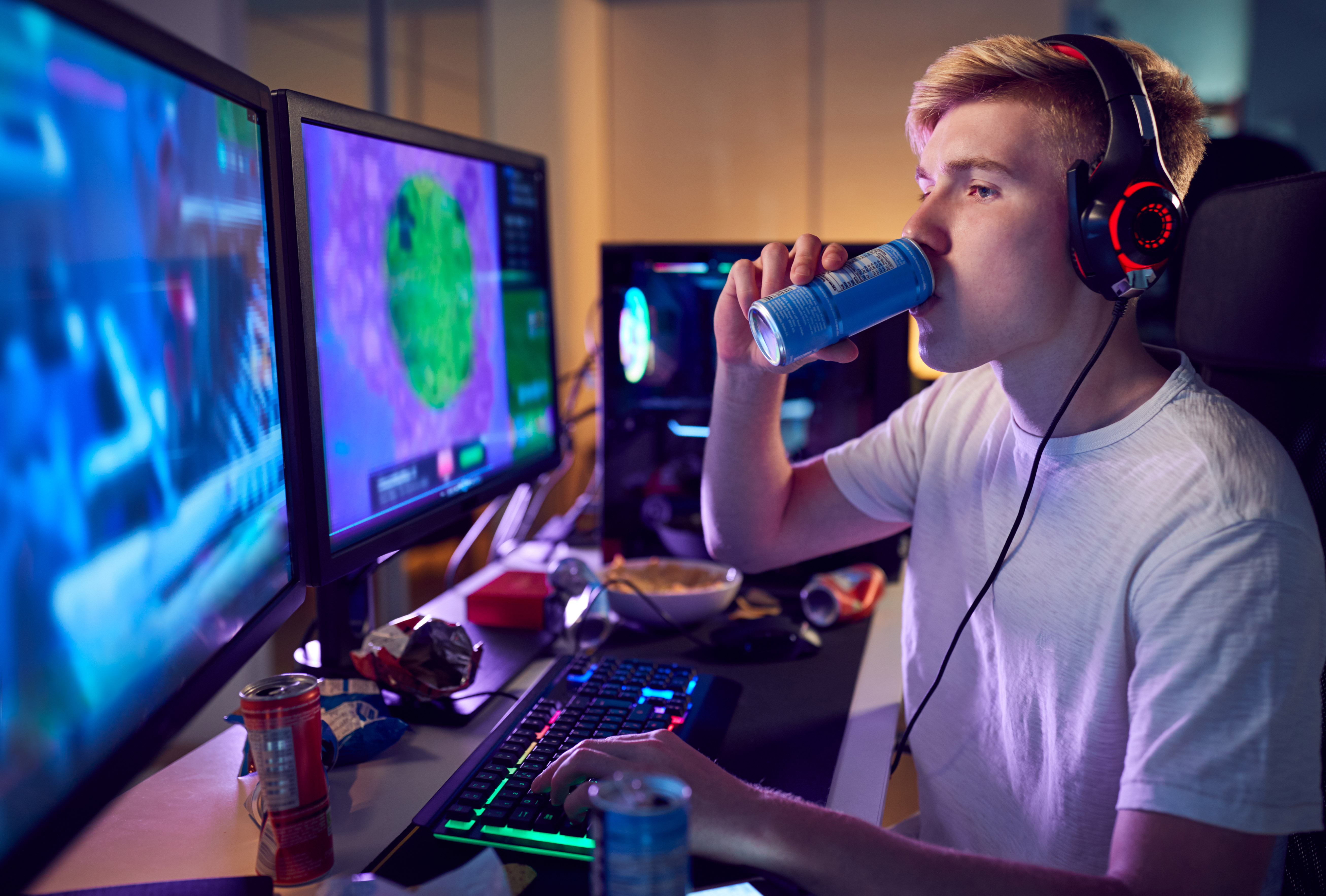 young man sits at computer surrounded by energy drink cans and chip bag wrappers. He drinks from a can while typing with his other hand