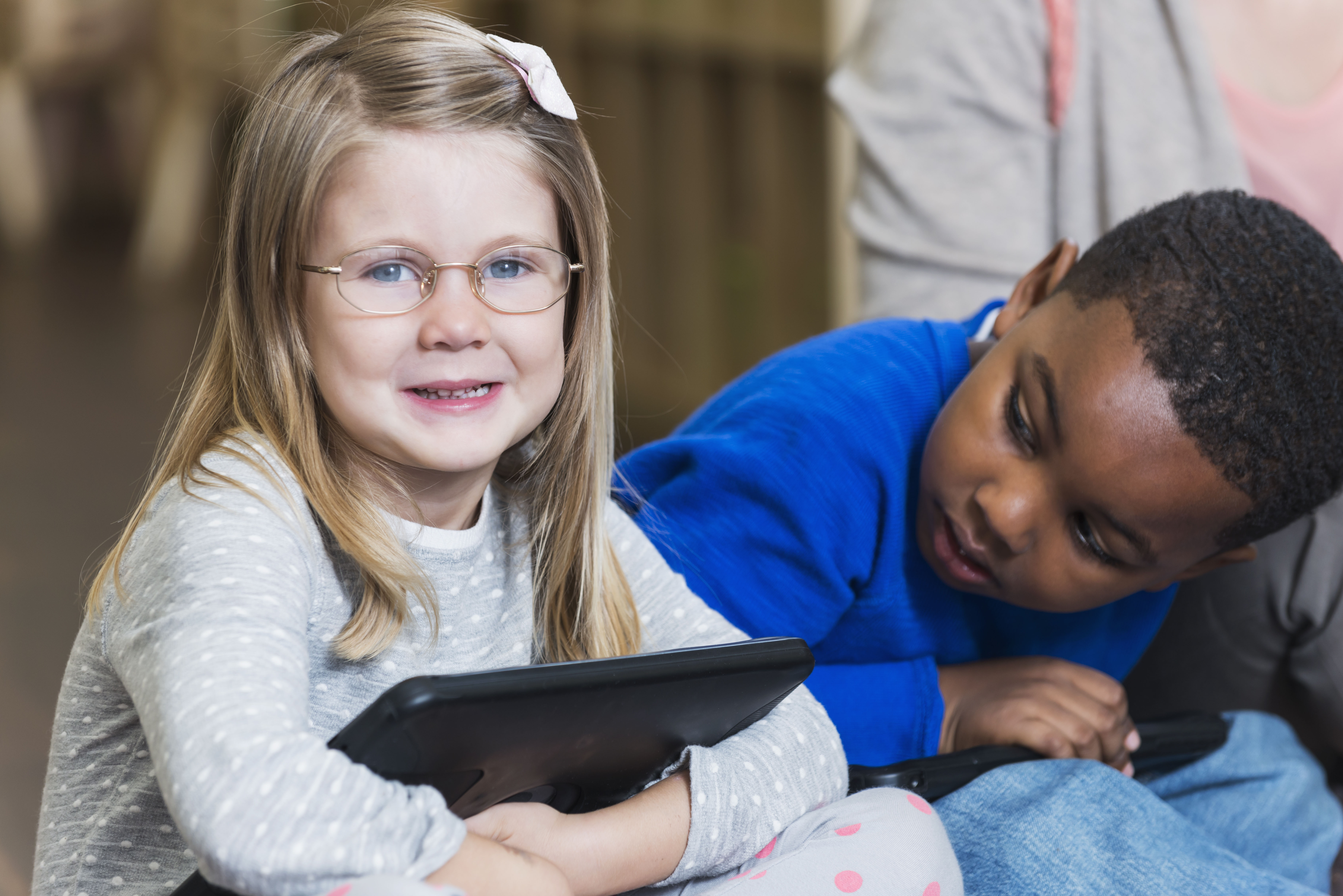 Two preschool students sit together. A White girl holds an iPad while a Black boy looks at her. 