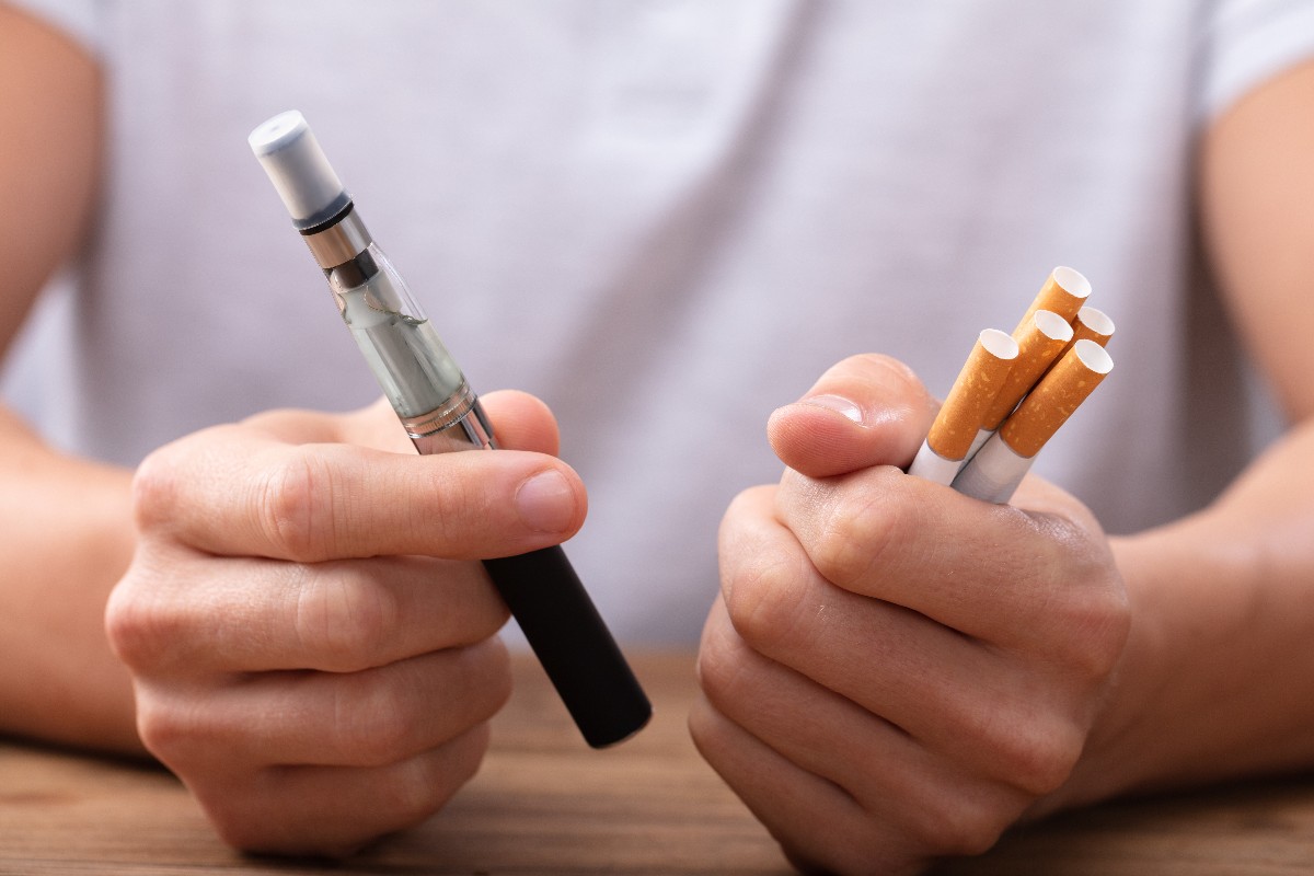 A man holds multiple cigarettes in his left hand and an electronic cigarette in his right hand. 