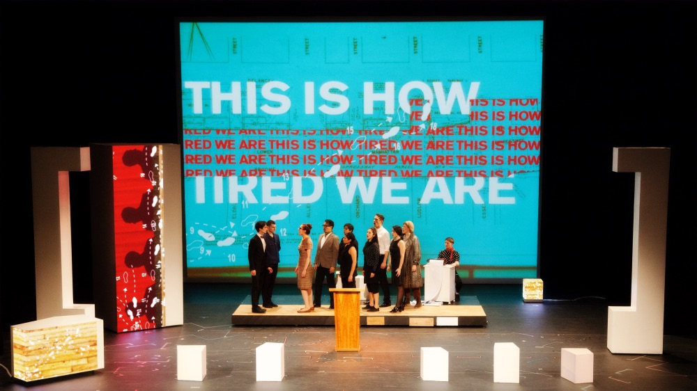 A group of people stand on a platform in front of a screen that reads "This is how tired we are."