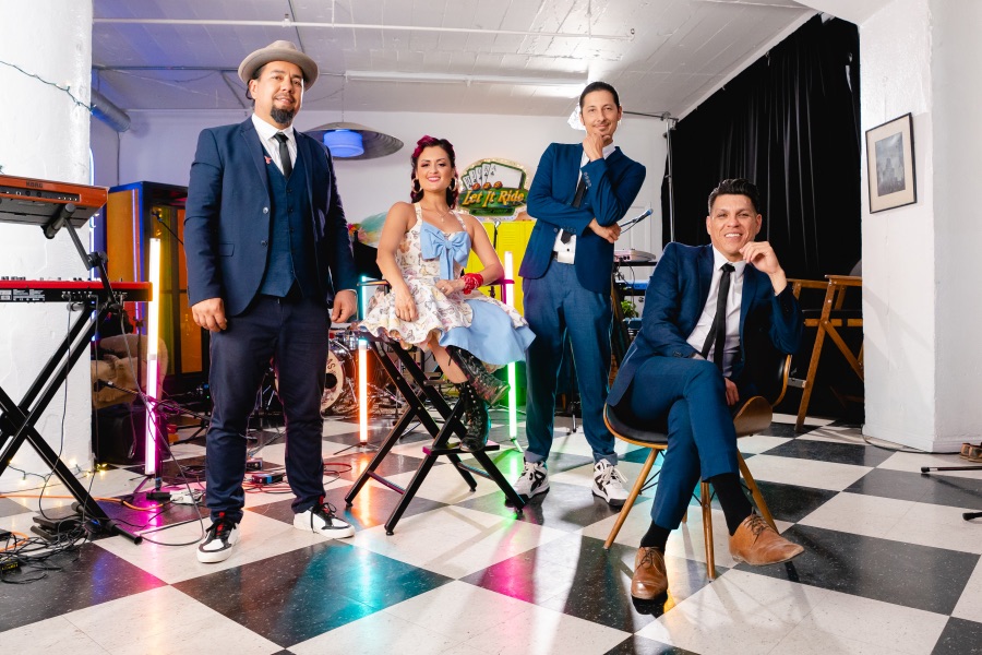 Four people wearing dapper formal clothing pose in a room next to keyboards.