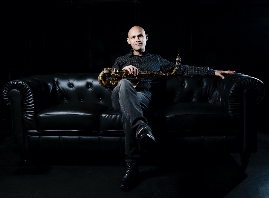 A bald man sits in the center of a tufted leather sofa as he holds a saxophone on his lap.