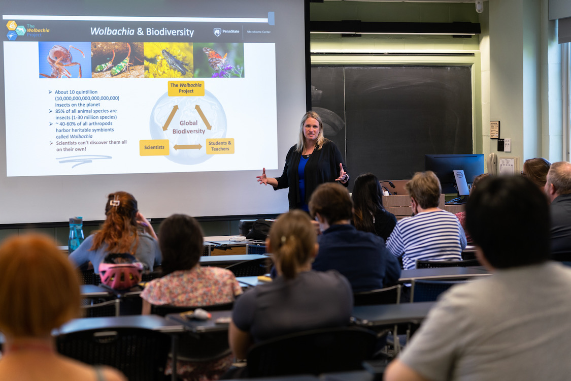 A woman speaks a to group in front of a screen featuring a power point presentation. 