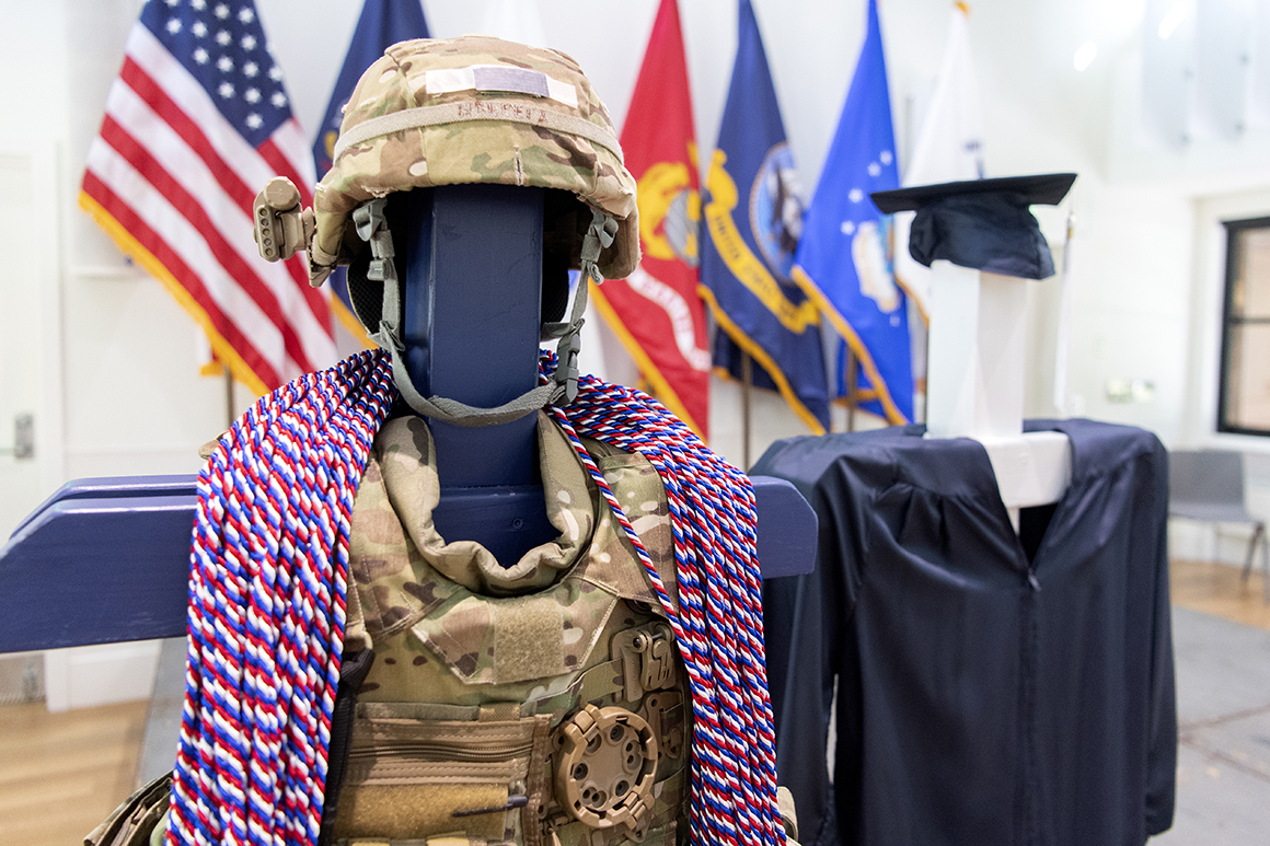 Camouflaged military vest and helmet with red, white and blue honor cords draped over next to blue undergraduate graduation cap and gown.