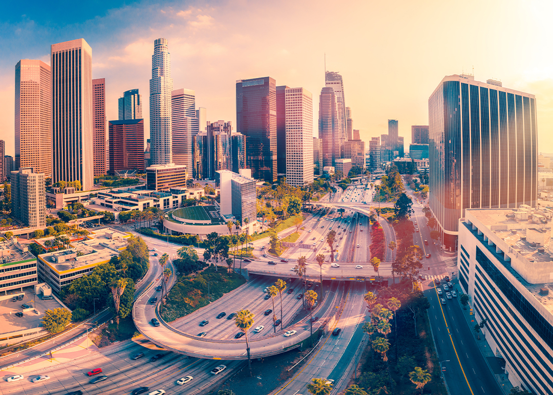 An image of the LA Skyline with the sun hitting it from behind with an overpass in the foreground. 