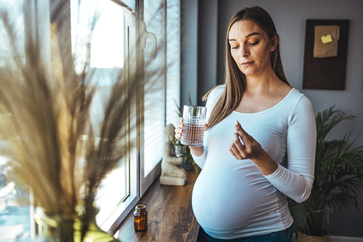 A pregnant woman stands by the window with a pill in her left hand and a drink in her right.