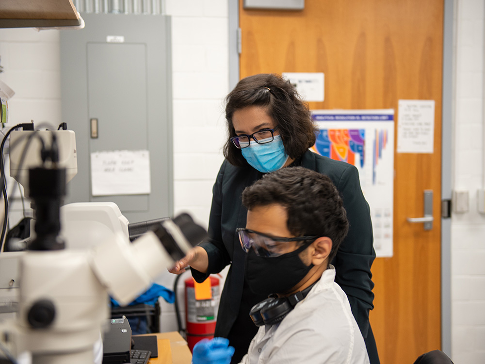 Dark-haired woman wearing black suit, glasses and mask stands next to dark-haired with mask and white lab coat seated at microscope in lab.