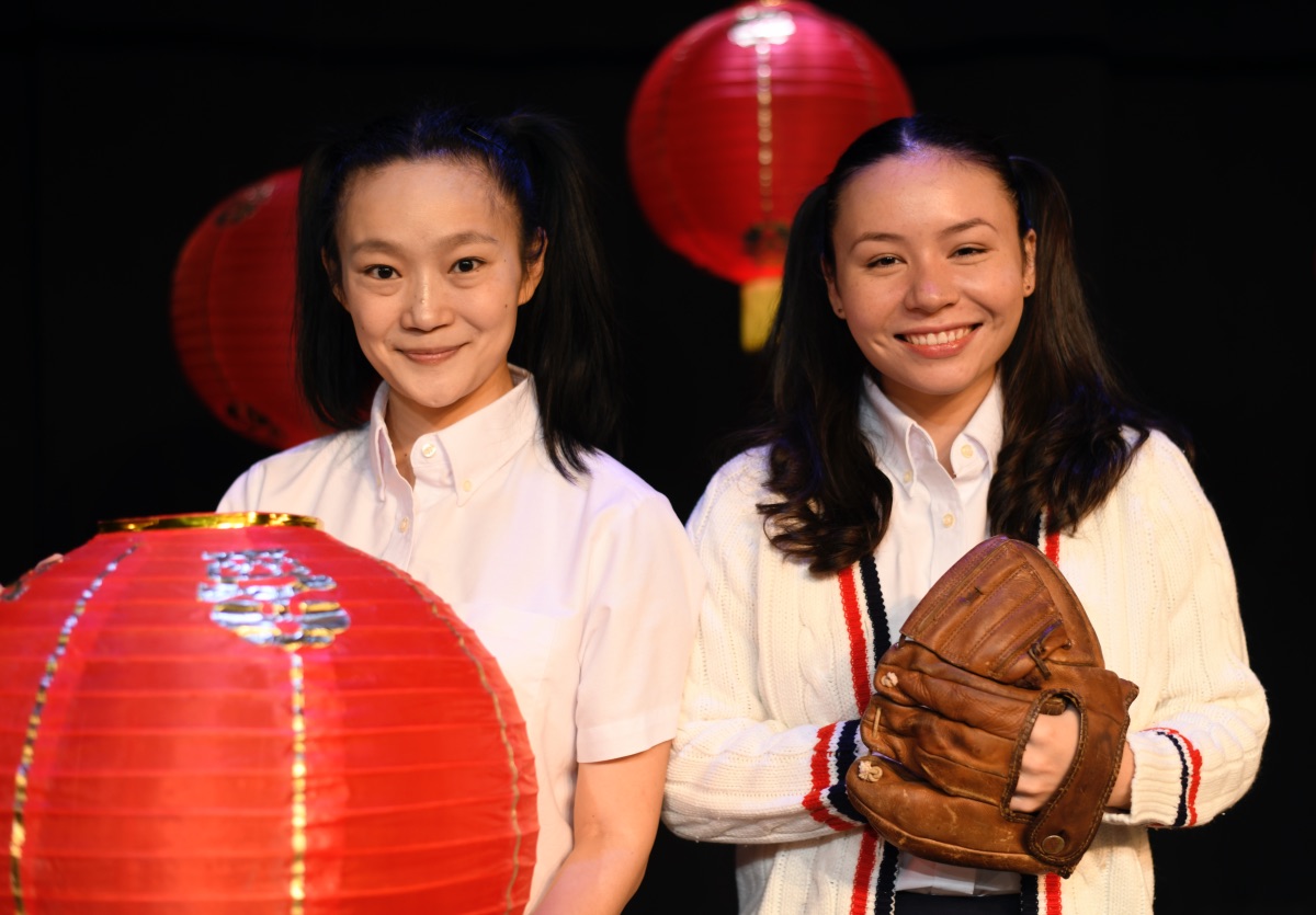 Two girls of Chinese heritage stand shoulder to shoulder, one holding a lantern, the other holding a baseballl glove.