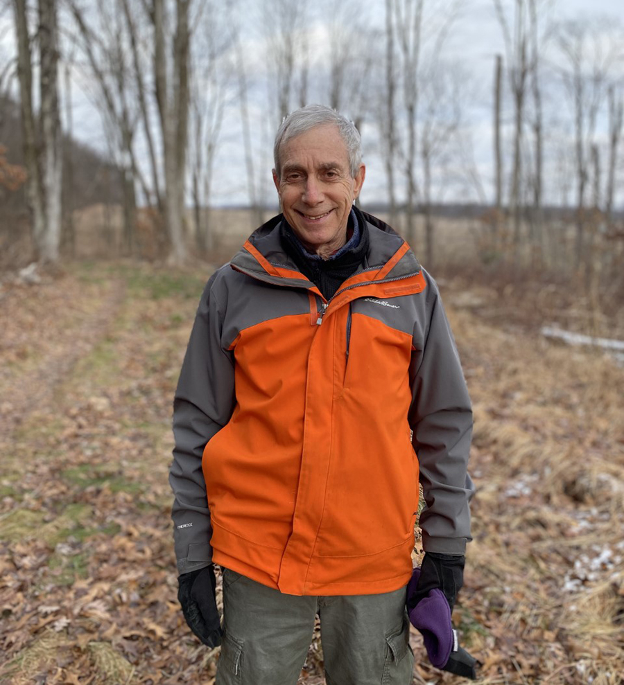 Retired professor Dave Eissenstat stands in a wooded area wearing an orange and gray jacket