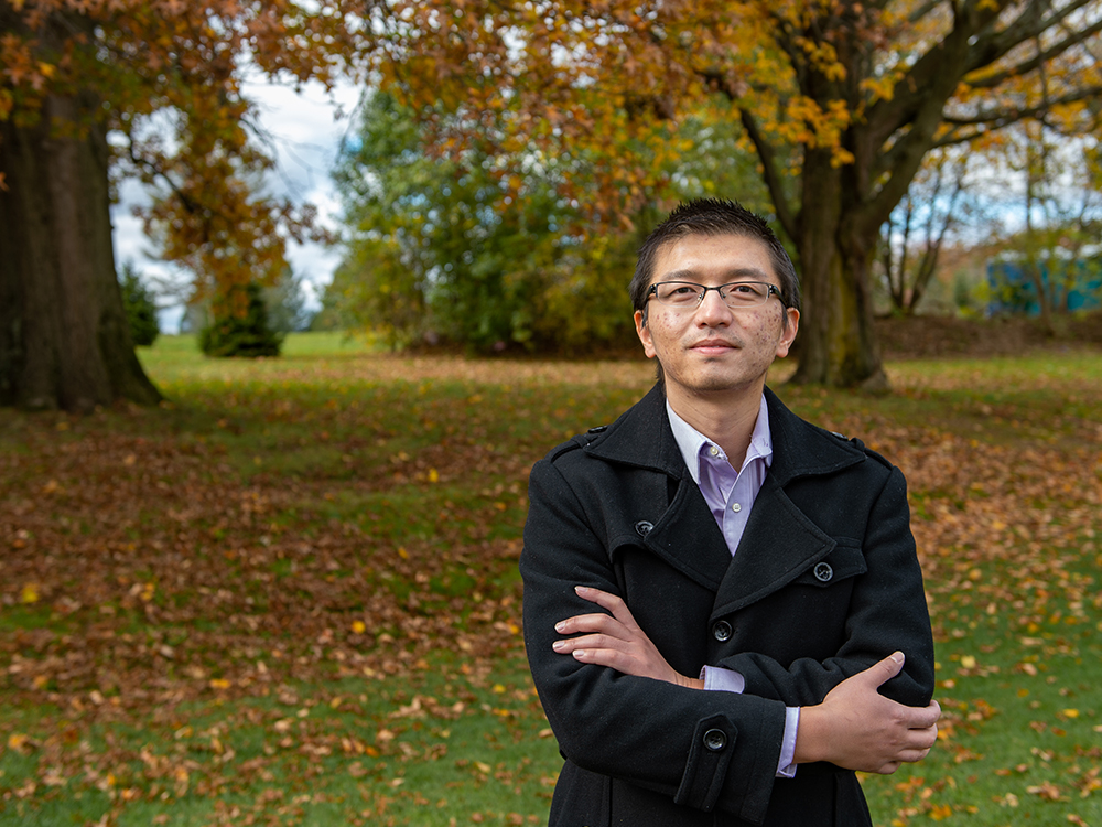 Dark-haired man wearing glasses and black wool coat stands outdoors with arms crossed.