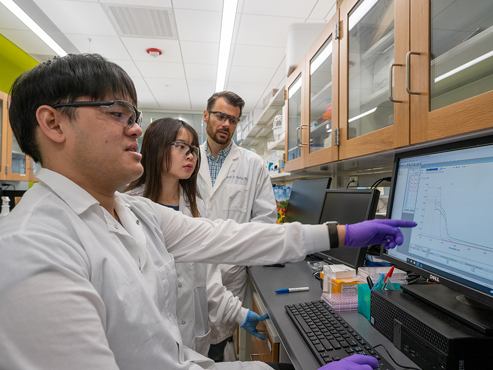 Three scientists in lab coats look at and point to a computer screen.