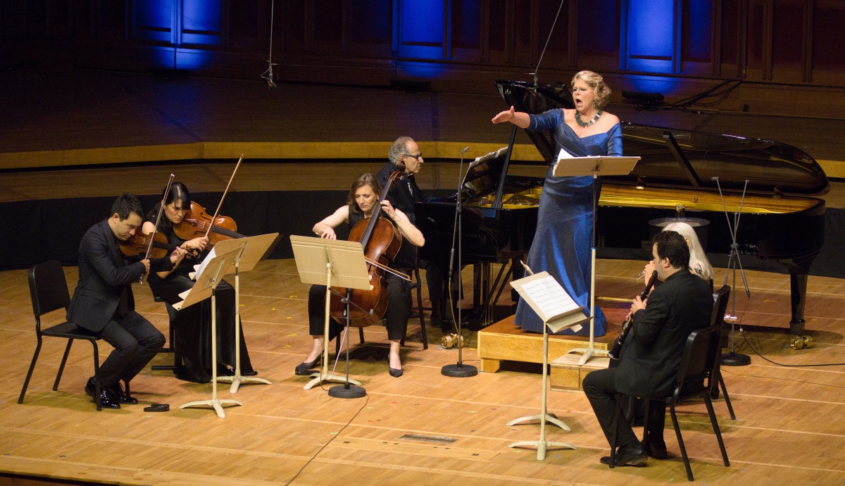 A classical music ensemble sits in a semi-circle while a vocalist wearing a formal gown sings.