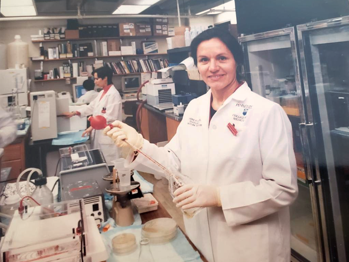 Joanna Floros stands in a laboratory wearing a white coat and holding a pipette.