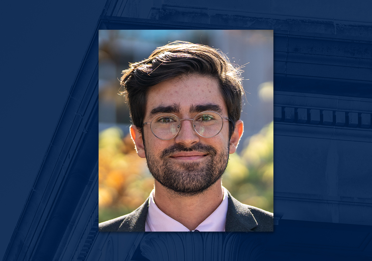 Man with dark hair, beard and glasses wears business suit and smiles in front of outdoor backdrop.