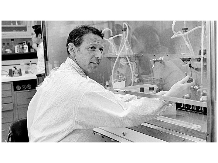 Paul Berg in the lab, working on an experiment using a lot of equipment behind glass. He's wearing a white lab coat and smiling at the camera.