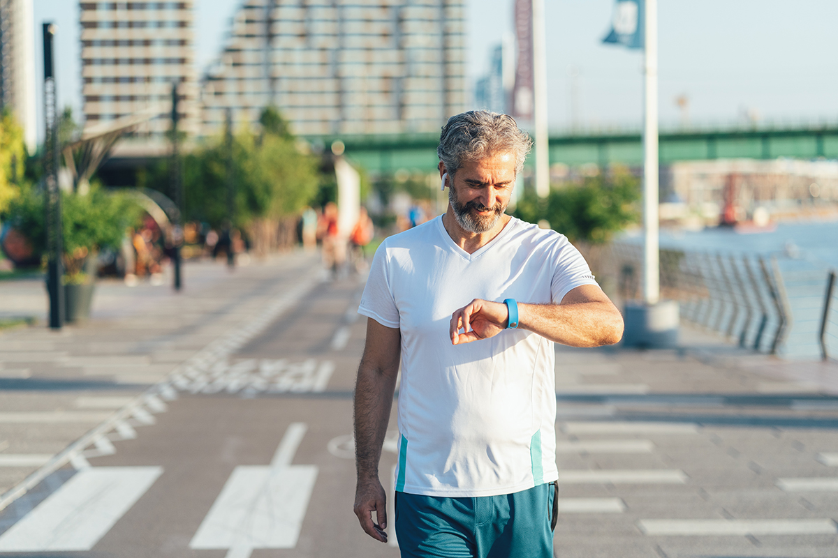 A middle-aged, bearded man wearing a short sleeve shirt walks along an urban waterfront while looking at his wearable device on his wrist. 