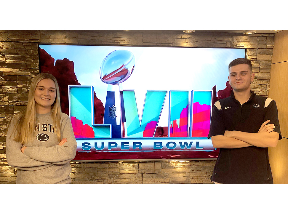 Two students with arms crossed standing on either side of a TV monitor