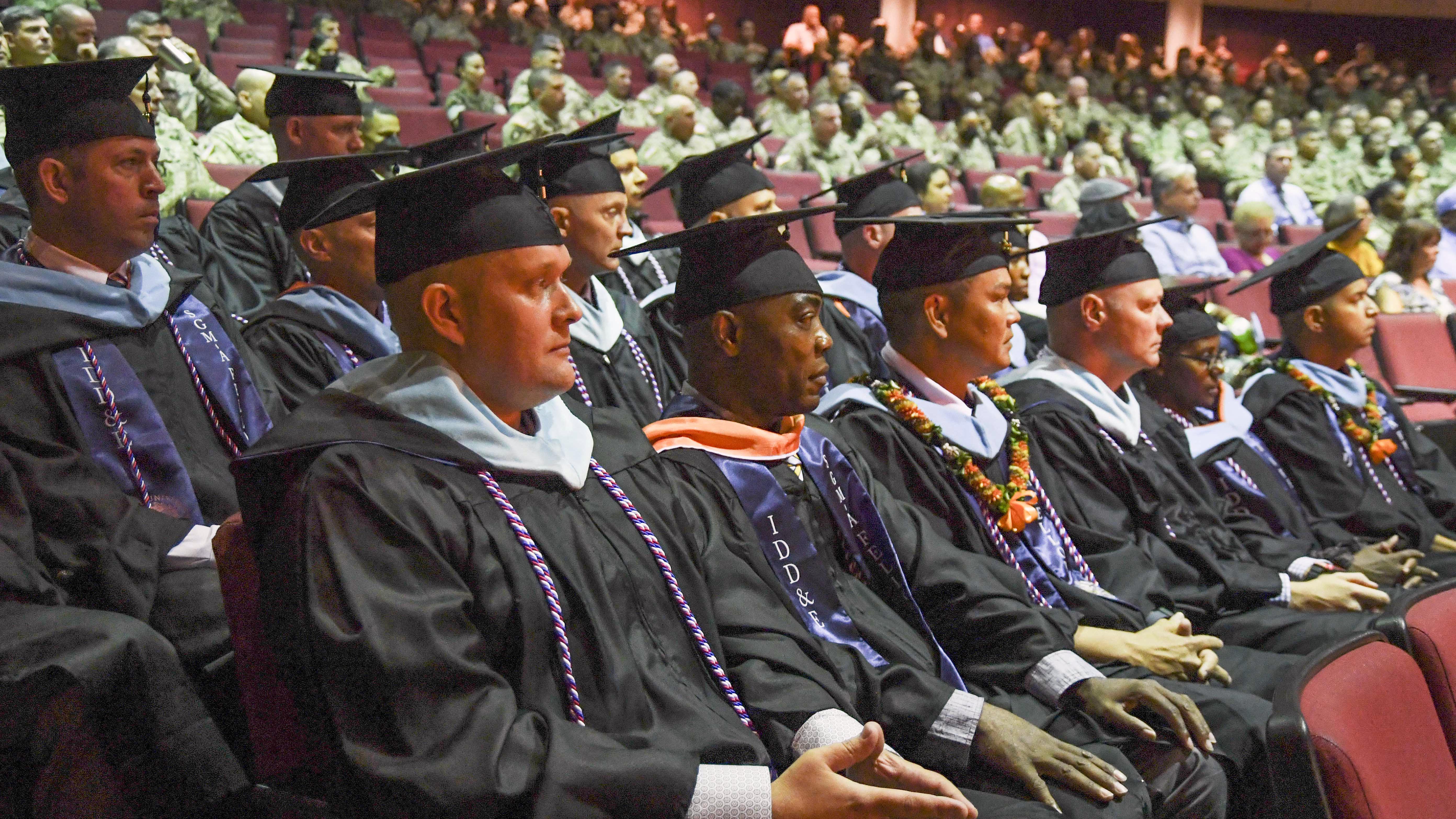 Soldiers wearing graduation caps and gowns and military cords around their necks sit in an auditorium