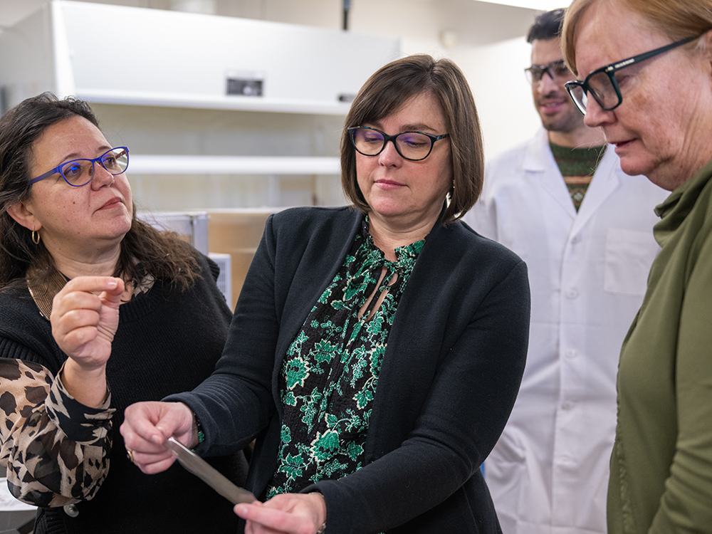 Three female researchers wearing business attire examine material in lab setting.