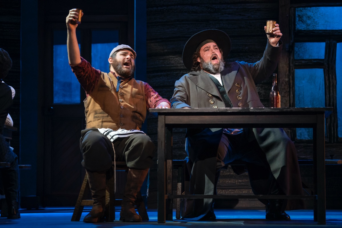 Two husky-figured Jewish men sit at a wooden table and lift their drinks in a toast.