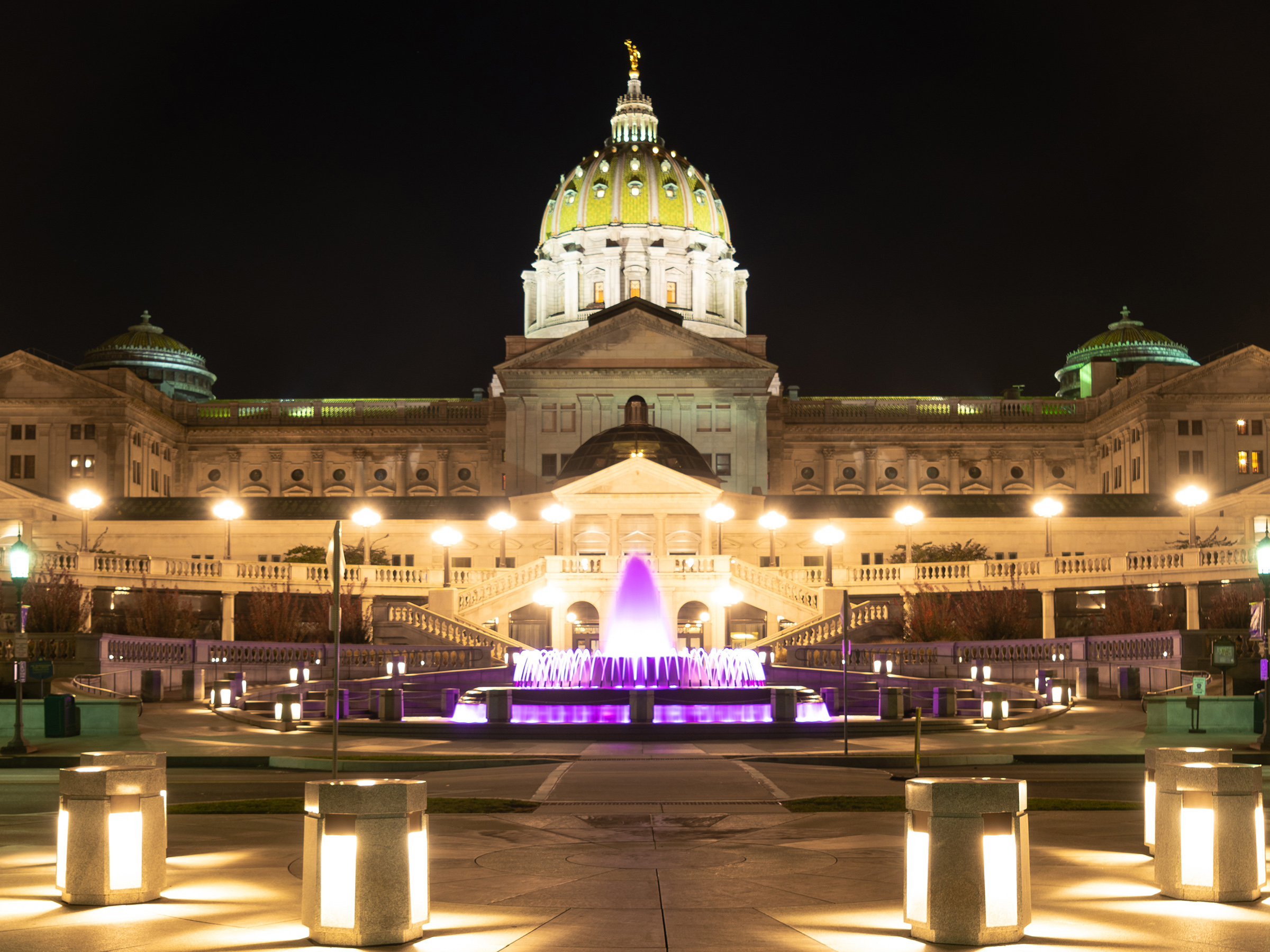 Exterior view of the Pennsylvania State Capitol Building at night with lights and a fountain illuminated in a purple light