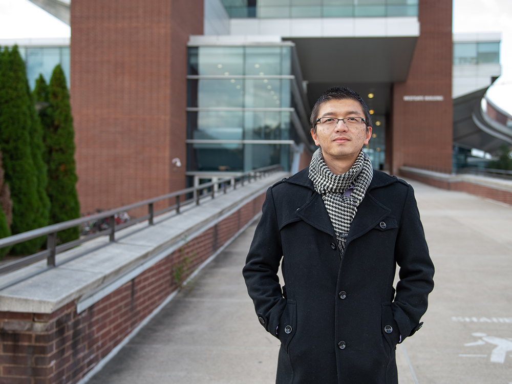 Dark-haired man wearing glasses, checked scarf, and black coat stands in front of red brick building.