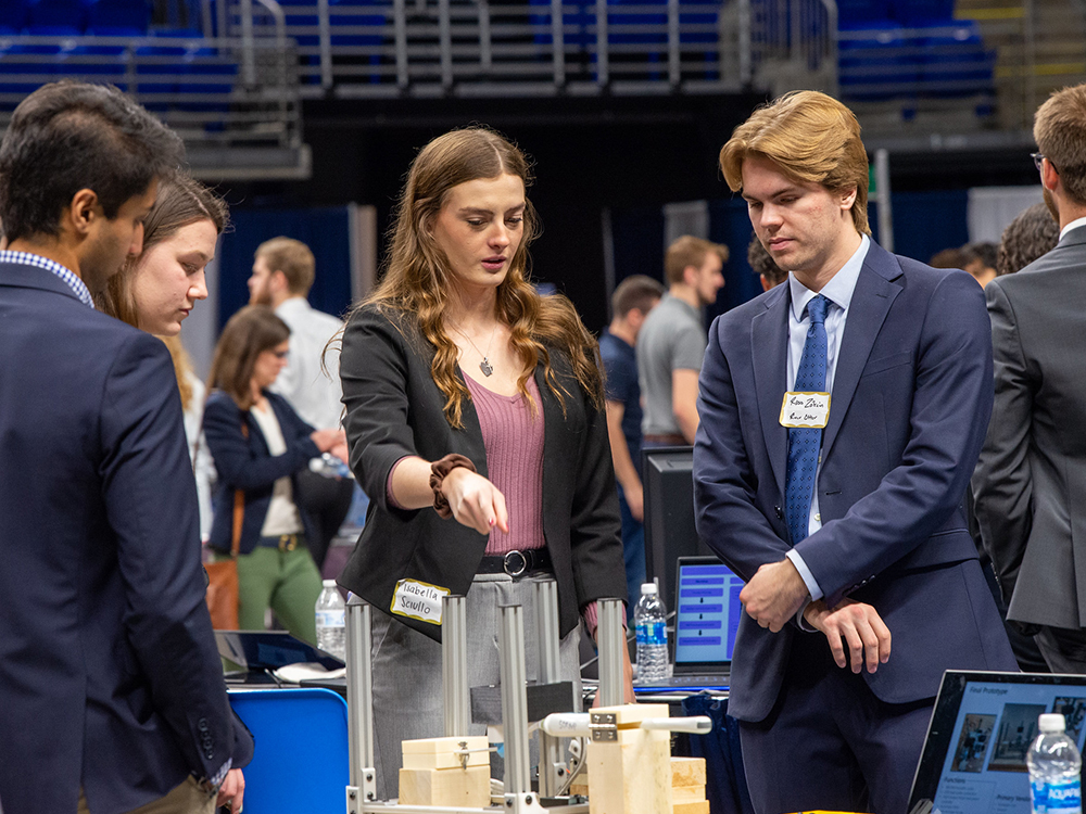 Young adults in business attire gather around an engineering exhibit.