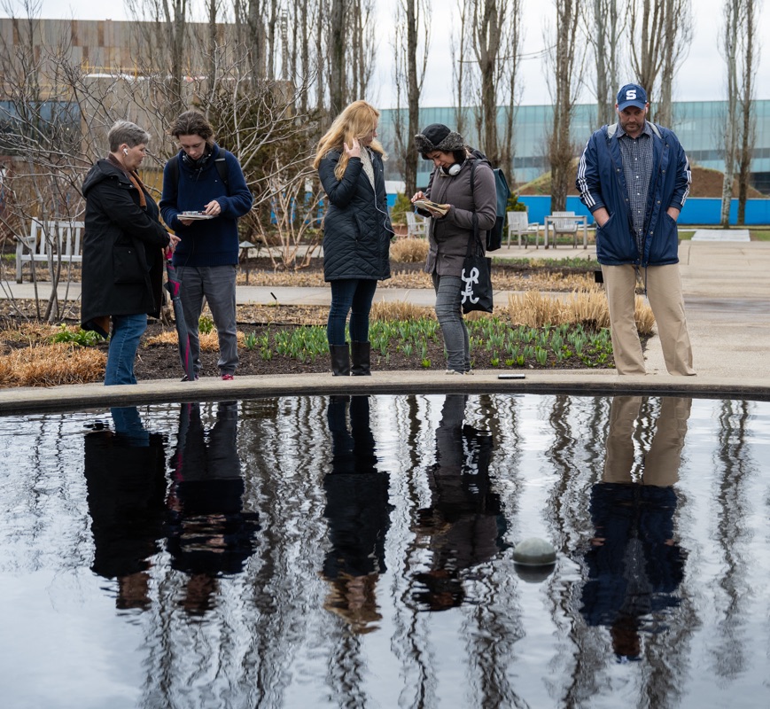 A group of five people pause in front of a reflecting pool as they listen to a prompt through headphones.