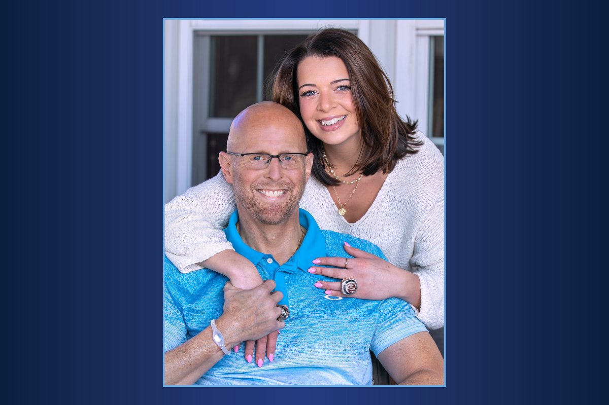 Sydney Harris, who has shoulder-length hair and wears her dad’s World Series Championship ring on her left hand, smiles as she encircles him in a hug. Doug, who is bald, is seated in front of Sydney and has a short beard, glasses and big smile on his face.