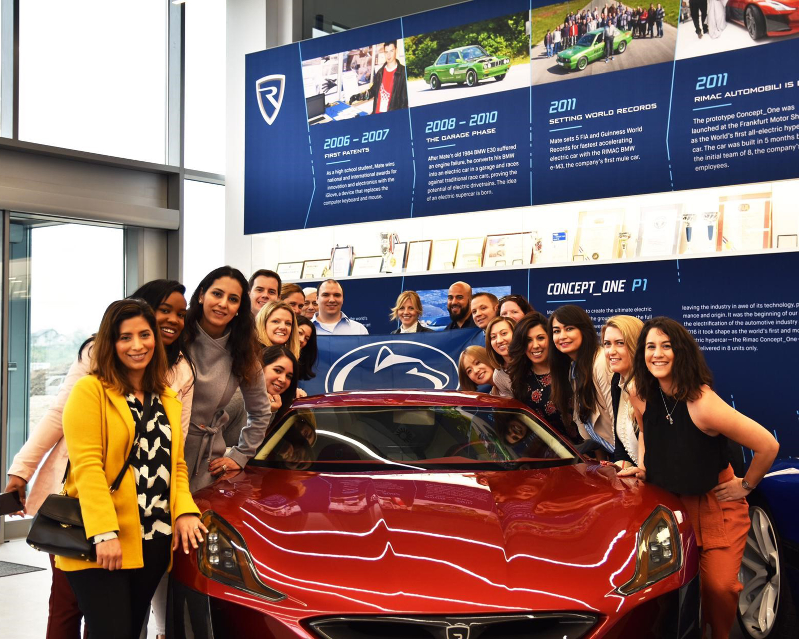 A group of people pose with a Penn State flag in front of a car.