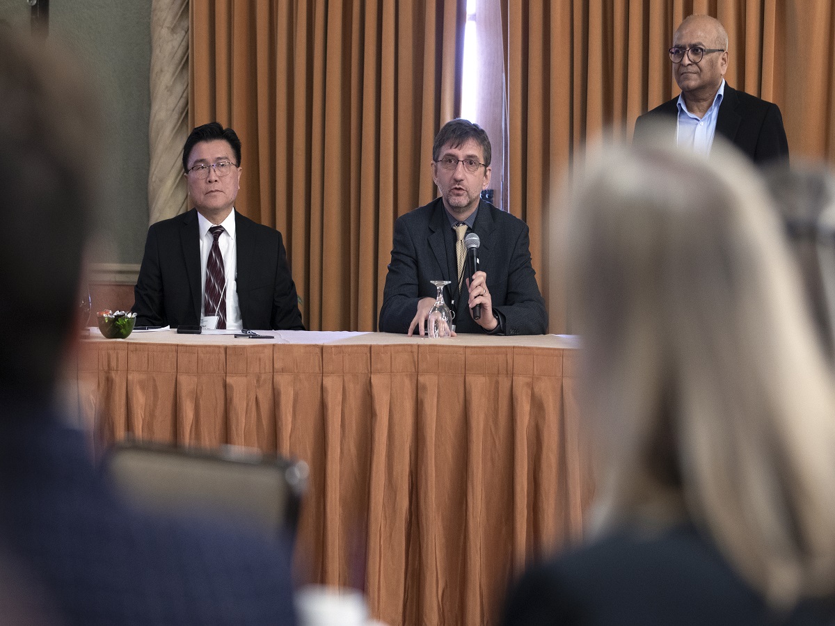 Three men are positioned behind a table while giving a presentation to a crowd. 