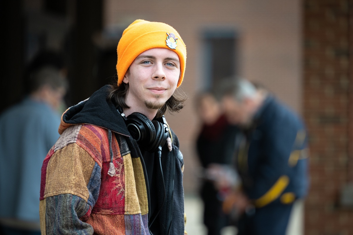 A young man wears a knit cap, a patchwork jacquard jacket and smiles as large headphones hang from his neck.