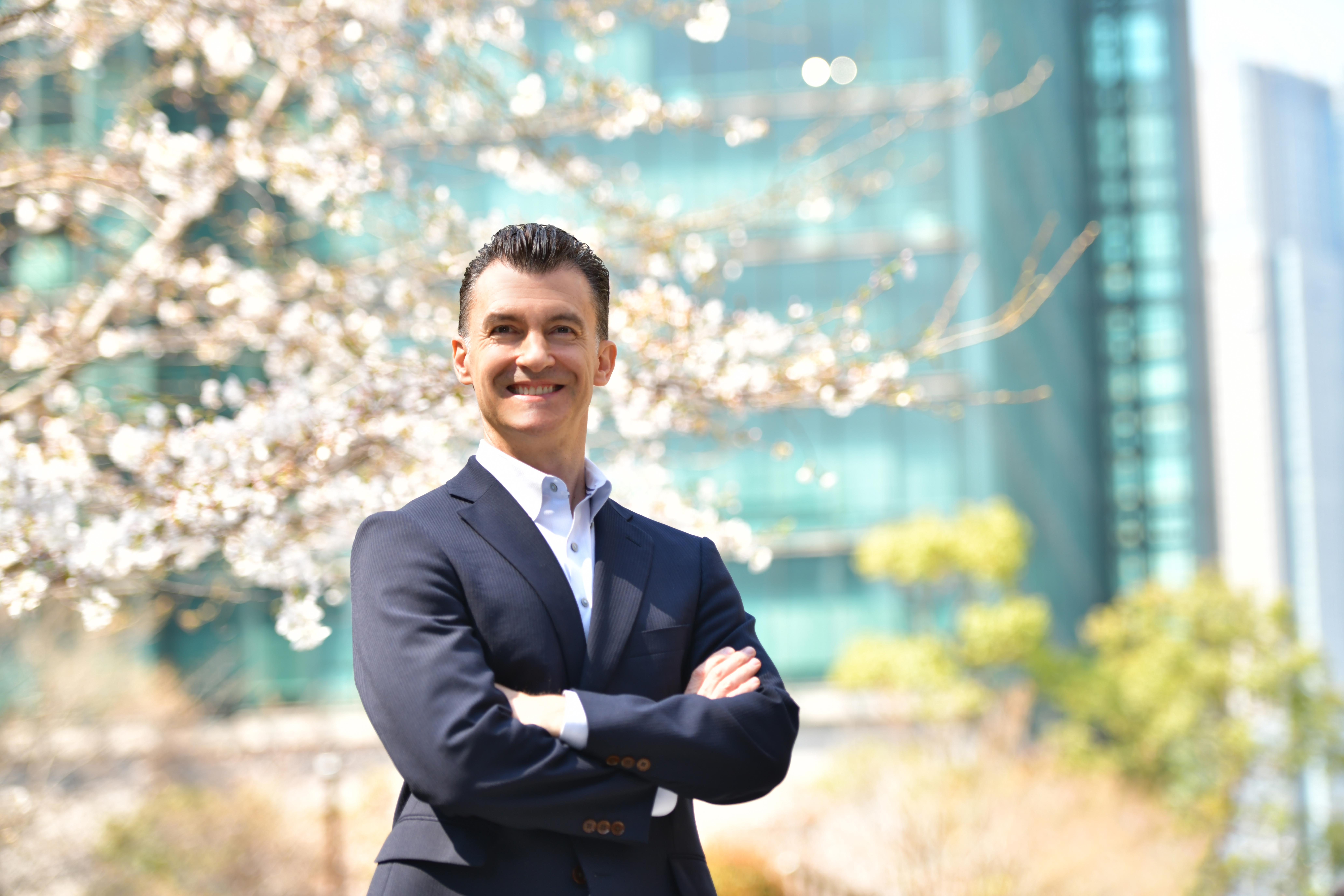 A man in a business suit, with his arms crossed, smiling for the camera, outside on a beautiful, sunny day