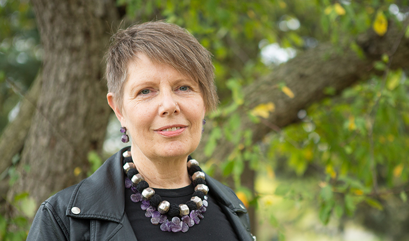 Robin Bower is pictured outdoors with trees in the back ground. She is wearing a black shirt, a chunky necklace and has short hair with side-swept bangs.