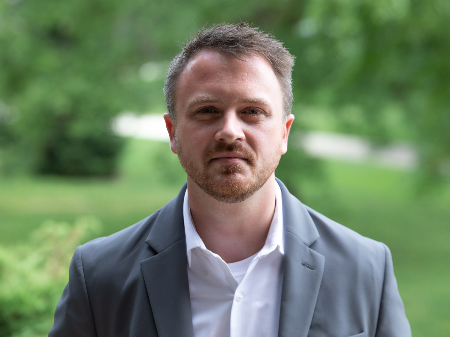 bearded man in sports jacket and white button-down shirt stands in front of green outdoor background