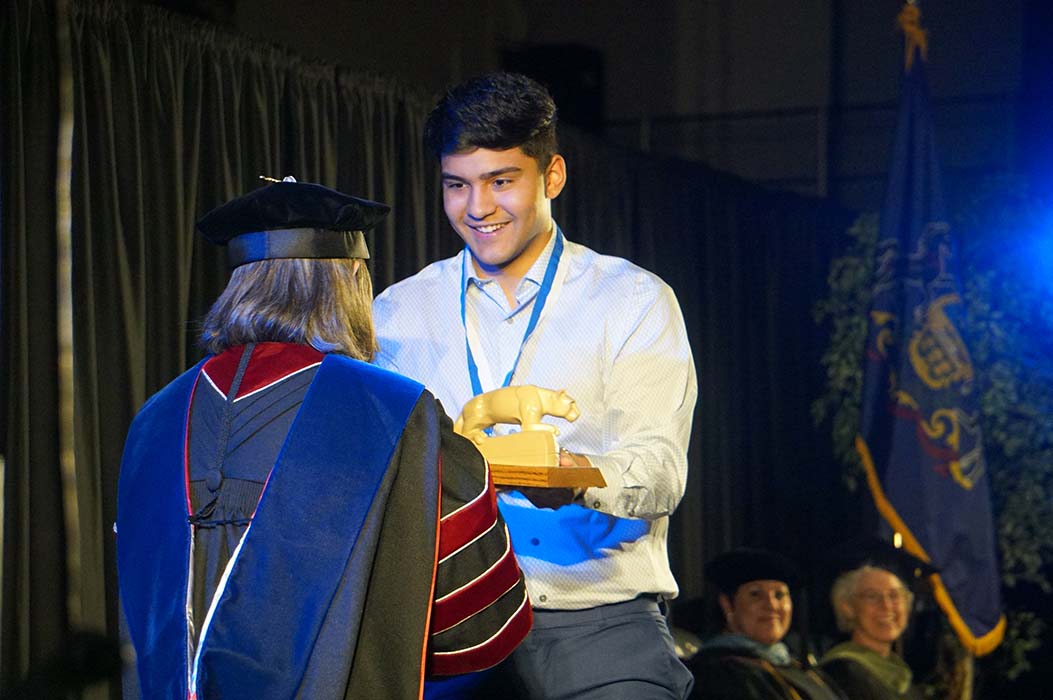 Male student in white dress shirt receiving a lion statue on a stage.