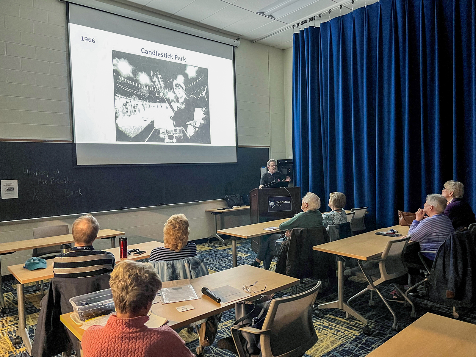 Back view of a classroom of older people at desks and a photo on the screen with instructor at front