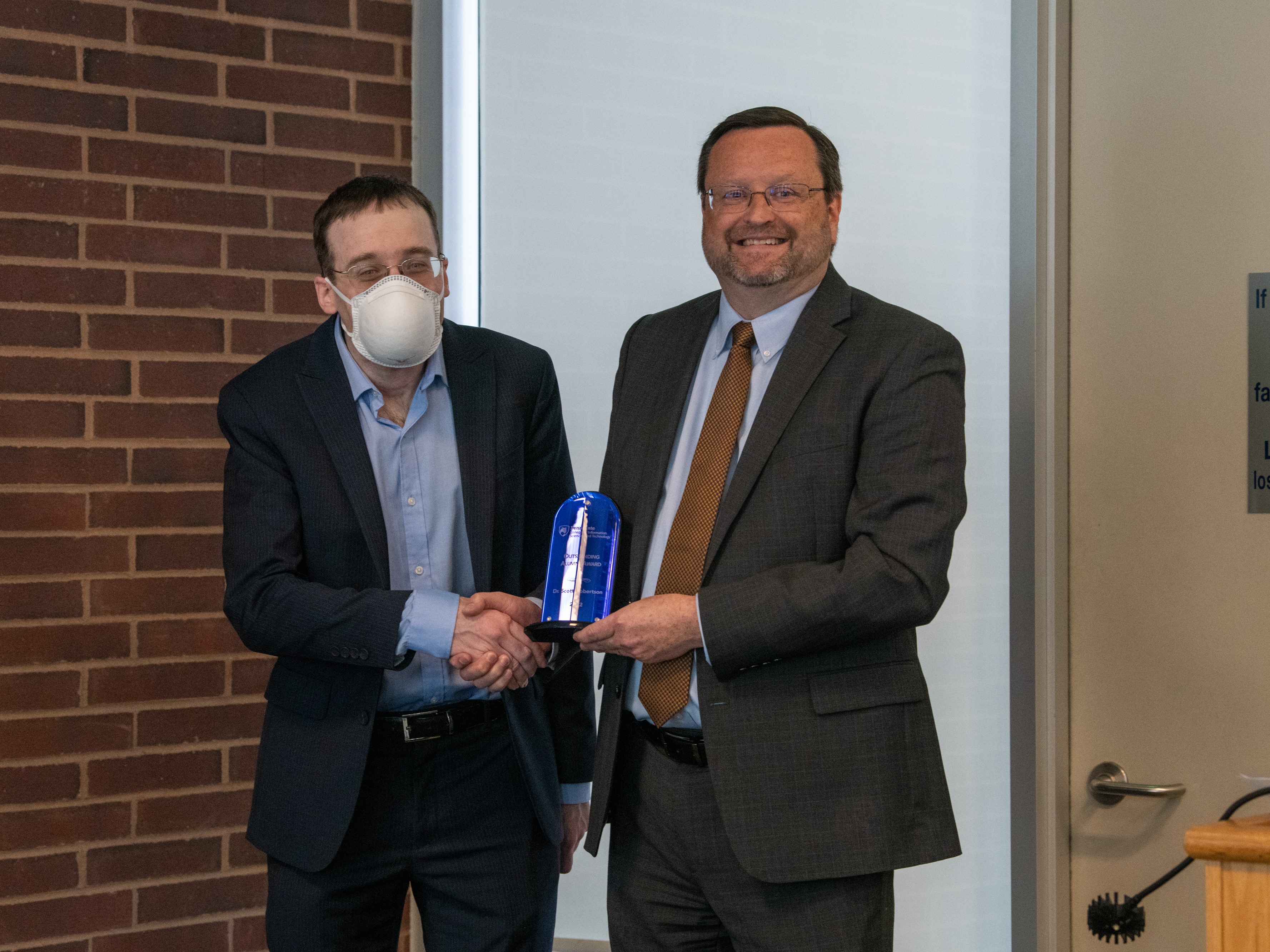 Man wearing suit and mask shakes hands with smiling man wearing glasses