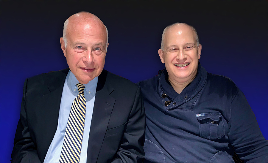 Photo of two seated men, one on left in coat and tie, one on right in a blue pullover