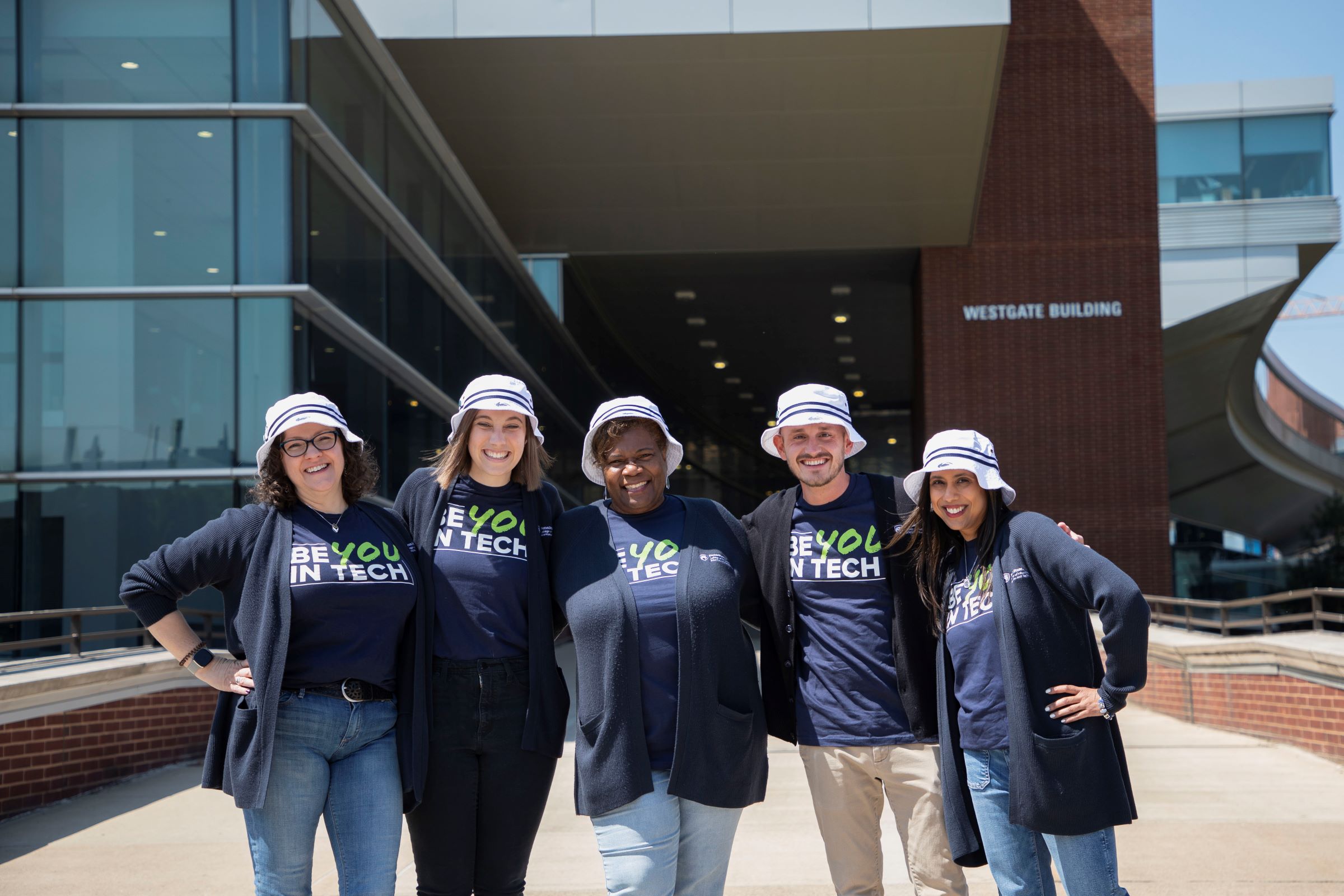 A row of five people in matching hats and shirts. A glass and brick building in the background.