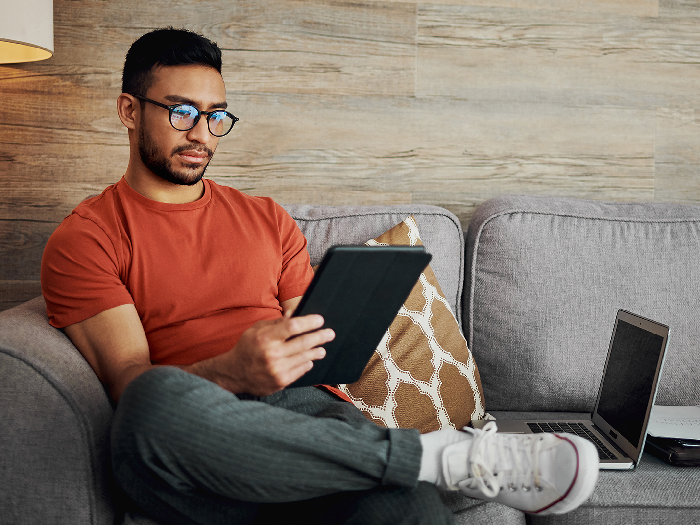 A man wearing glasses reads from a digital tablet, relaxing on a couch.