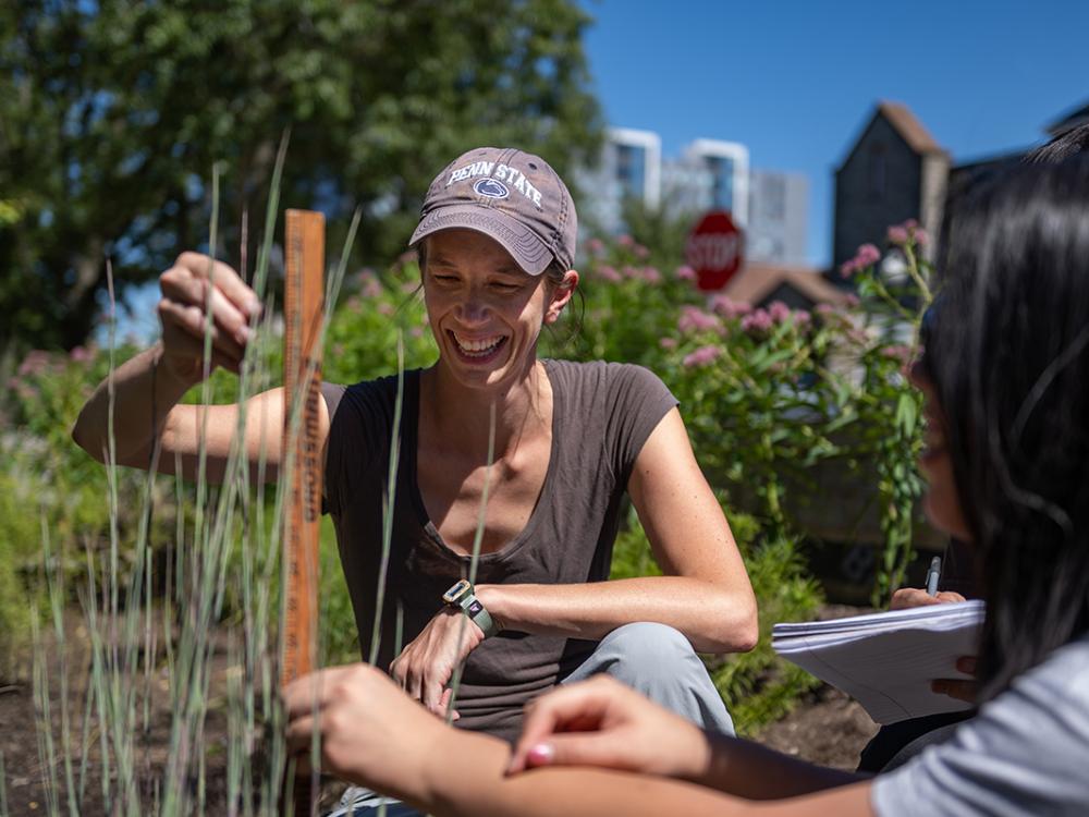 Lauren McPhillips conducting outdoor research by using a ruler to measure vegetation. She is smiling, wearing a baseball cap, and with another person who is helping her.