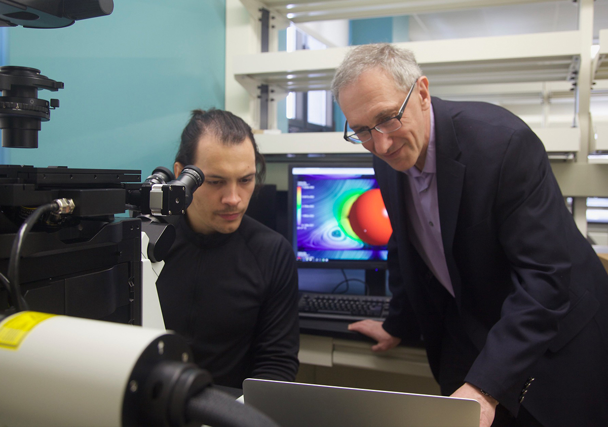 Two people sit in front of a microscope and look a a laptop