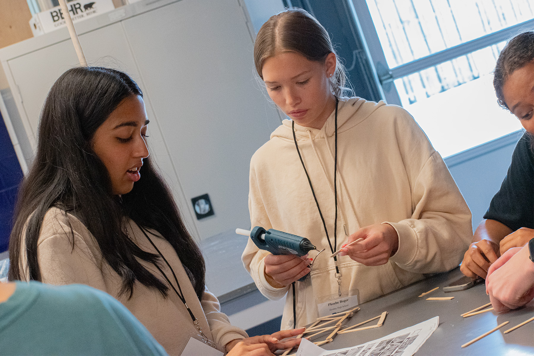 Photo of two students looking at instructions for building a bridge from popsicle sticks. One student holds a hot glue gun.