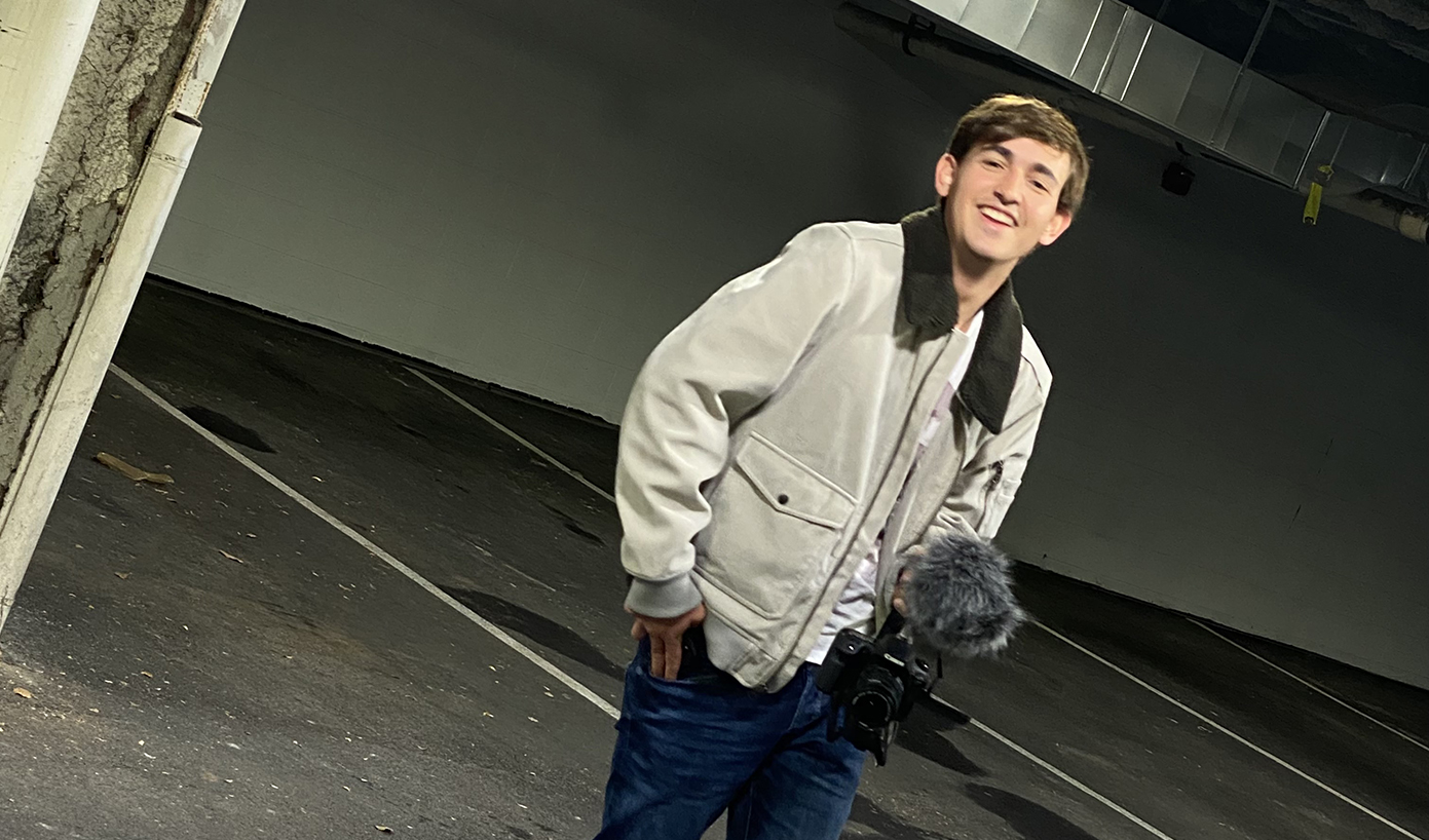 A smiling dark-haired student in a tan coat and jeans